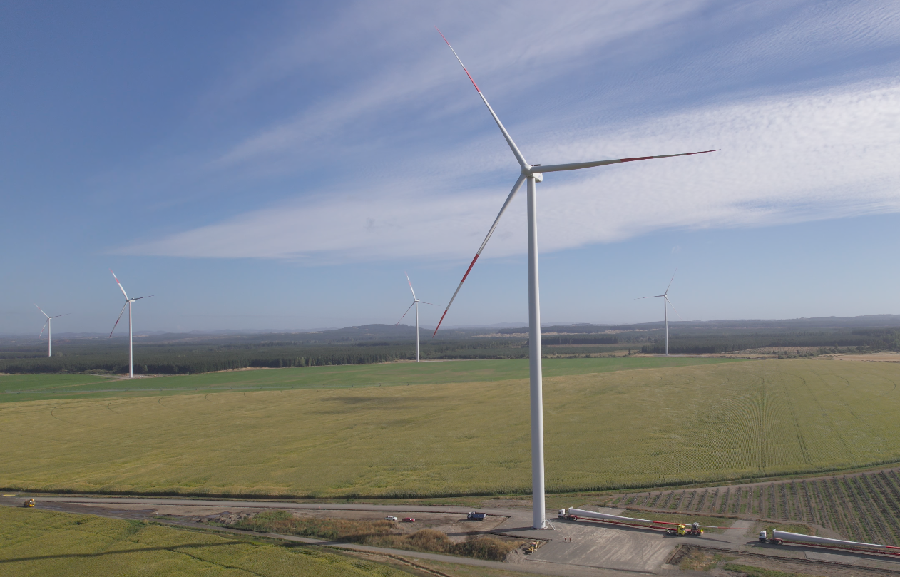 A landscape with several wind turbines in a vast field under a blue sky with light clouds. The turbines are tall with large blades, and there are vehicles and equipment at the base of the nearest turbine.