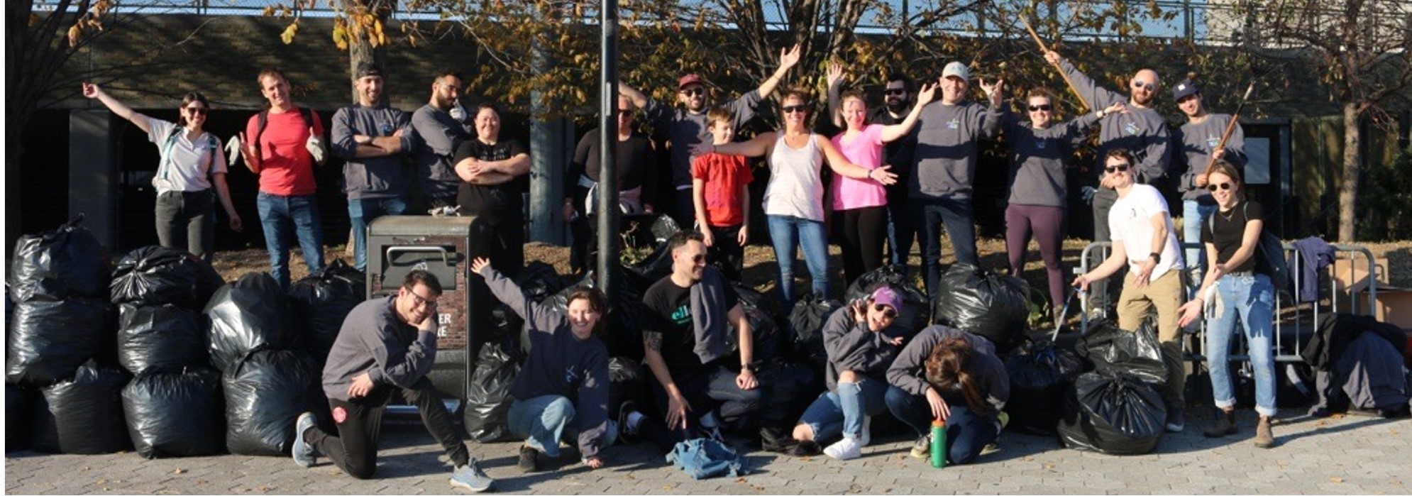 A group of people posing outdoors with garbage bags after a cleanup event. They are smiling and appear to be celebrating their efforts. Trees with autumn leaves are visible in the background.