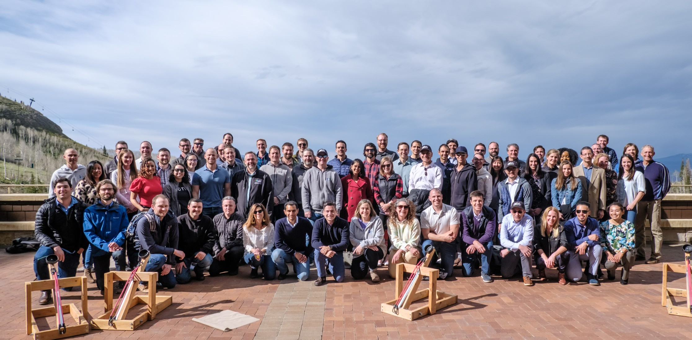 A large group of people posing for a photo outdoors on a brick patio with a mountainous landscape in the background. They are standing and kneeling in rows, with three wooden catapult-like structures in front.