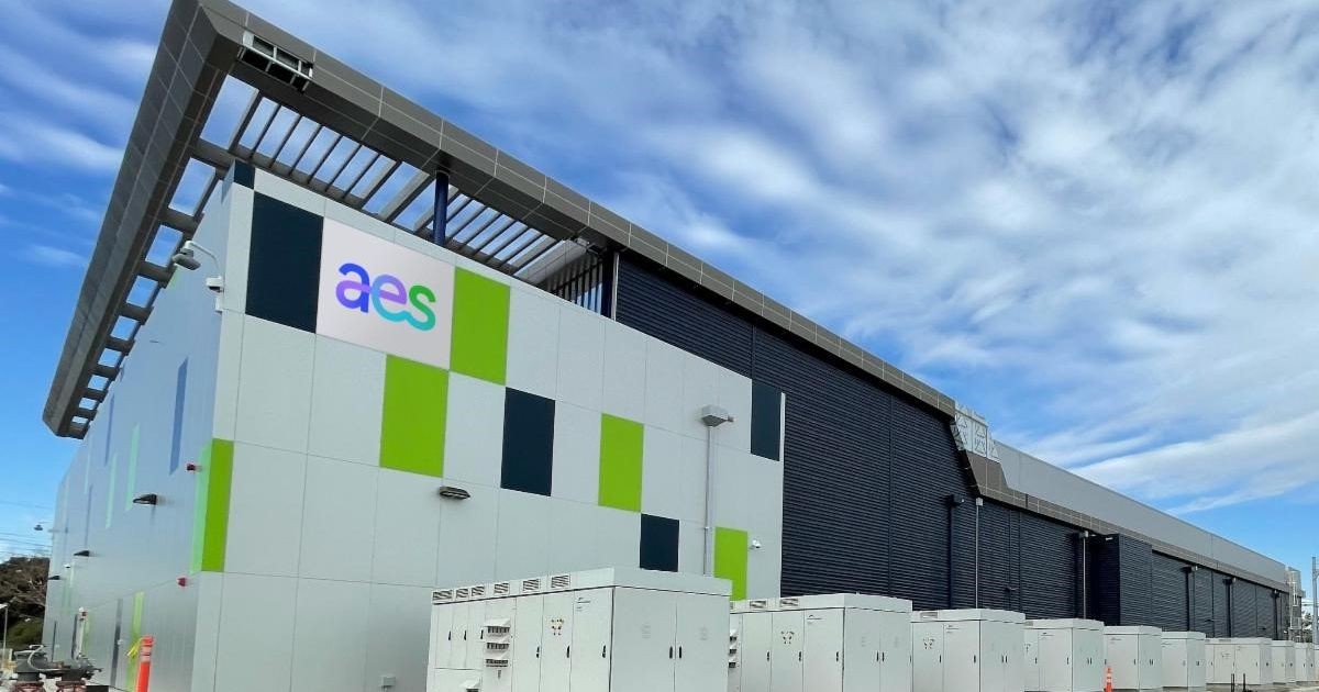 Modern industrial building with a large logo on the side, featuring green and dark panels. Several electrical units are visible in the foreground under a blue sky.