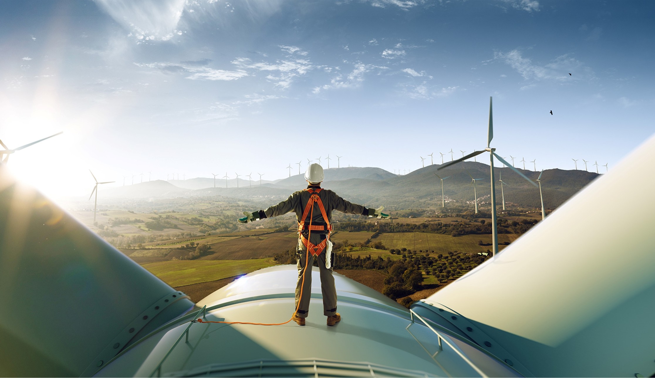 Person in safety gear stands on a wind turbine with arms outstretched, overlooking a landscape with multiple wind turbines and mountains in the background.