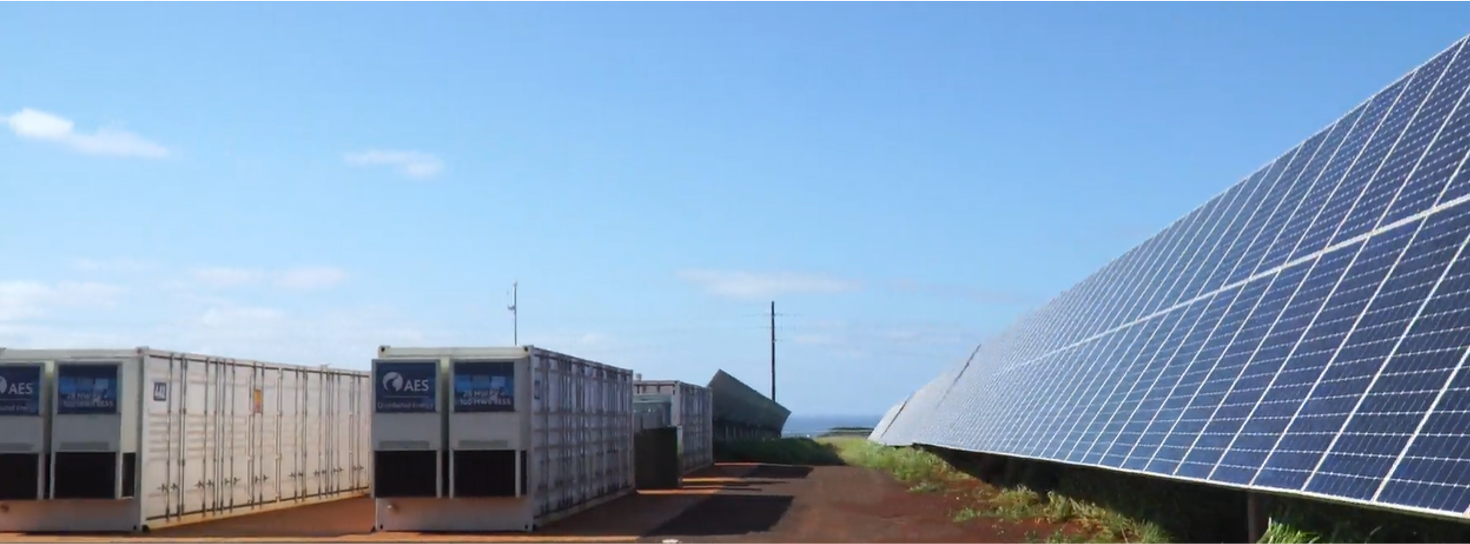 A solar power facility with large solar panels on the right and several white storage containers on the left under a clear blue sky.