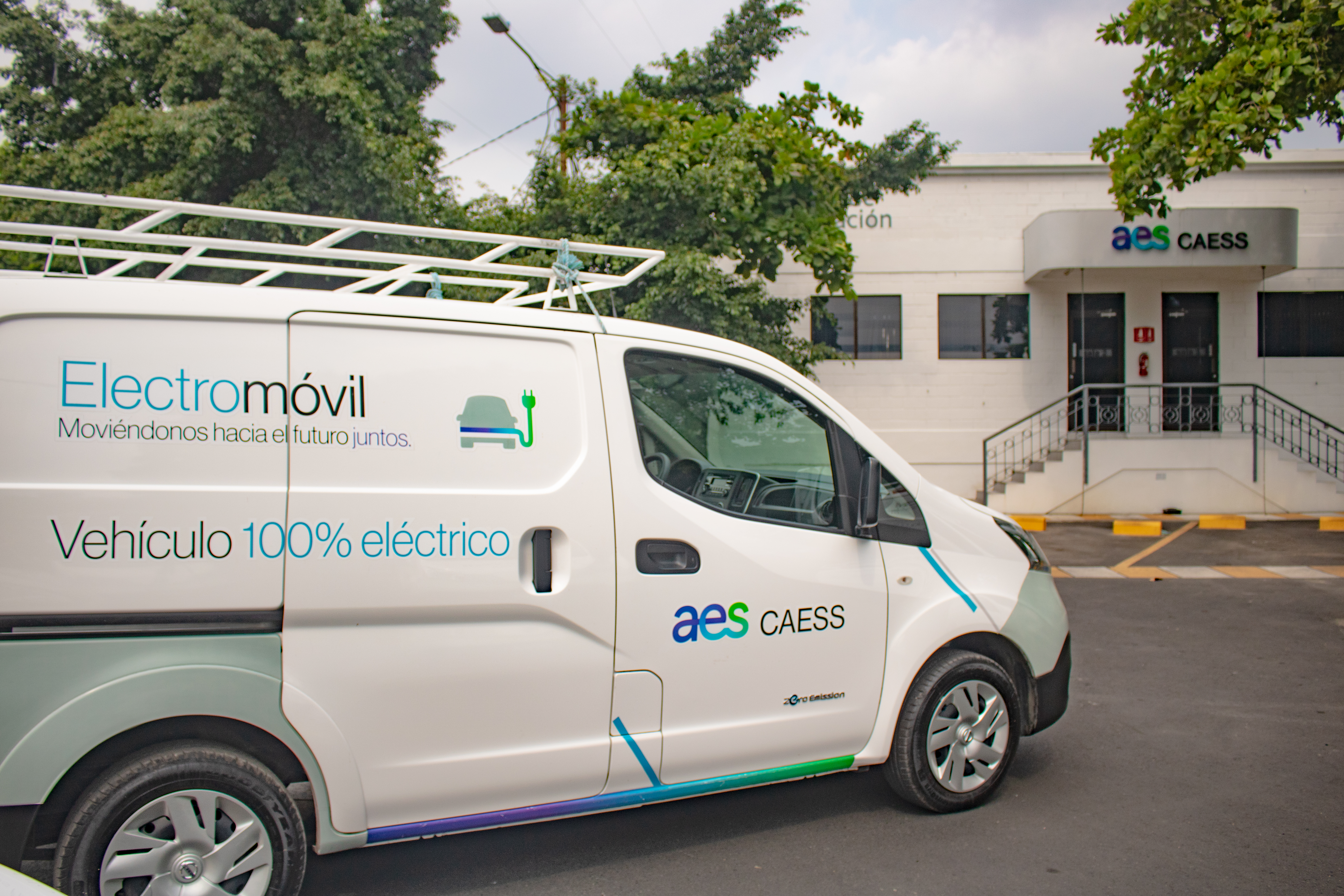 A white electric van with the text 'Electromóvil, Moviéndonos hacia el futuro juntos. Vehículo 100% eléctrico' and the logo 'aes CAESS' is parked in front of a building with the same logo. The van has a roof rack, and trees are visible in the background.