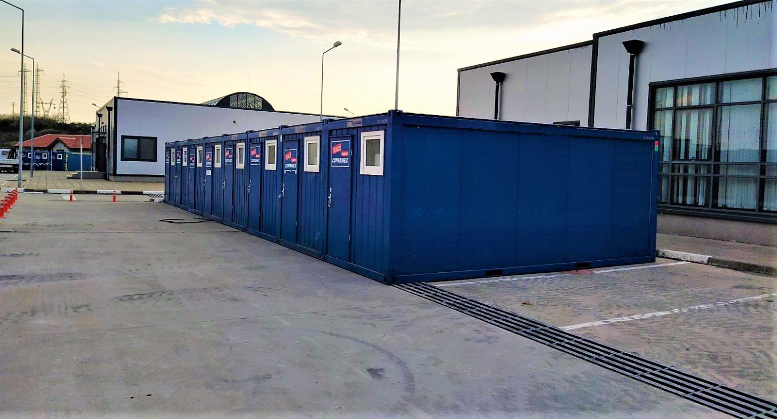 A row of blue shipping containers lined up outside a building with large windows. The containers have small windows and doors, and are placed on a paved area. Streetlights and power lines are visible in the background.