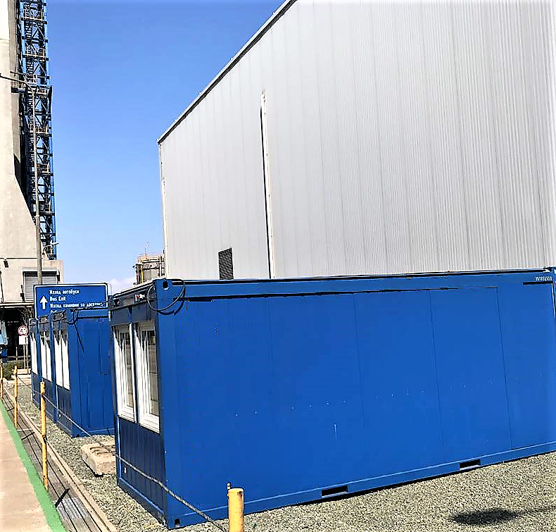A large industrial building with a corrugated metal wall is adjacent to two blue shipping containers with windows. A blue sign with white text and an arrow is visible near the containers. The ground is covered with gravel, and a yellow post is in the foreground.