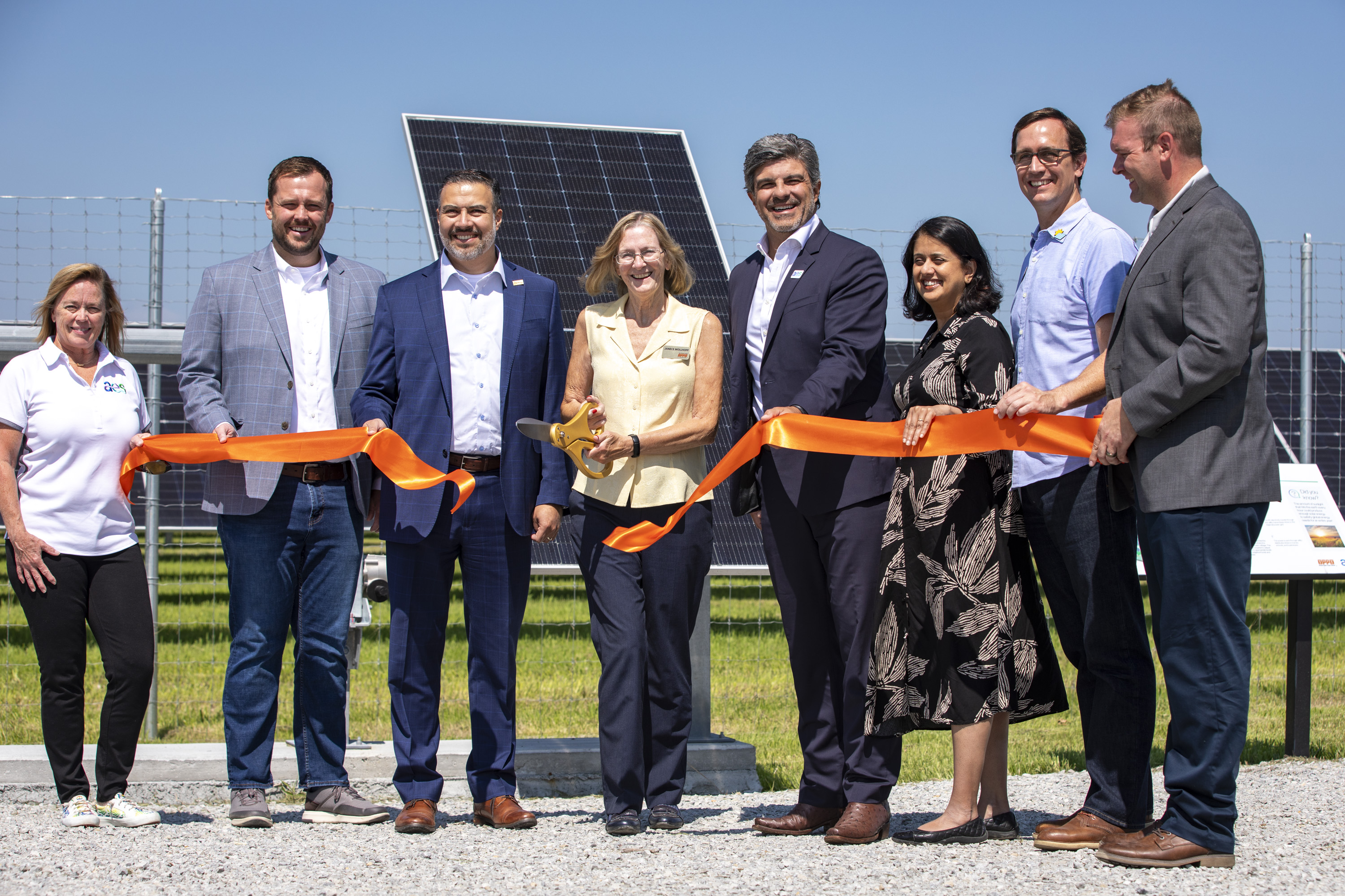 A group of people stand in front of solar panels, holding an orange ribbon at a ribbon-cutting ceremony. One person holds large scissors, ready to cut the ribbon. The event takes place outdoors under a clear blue sky.