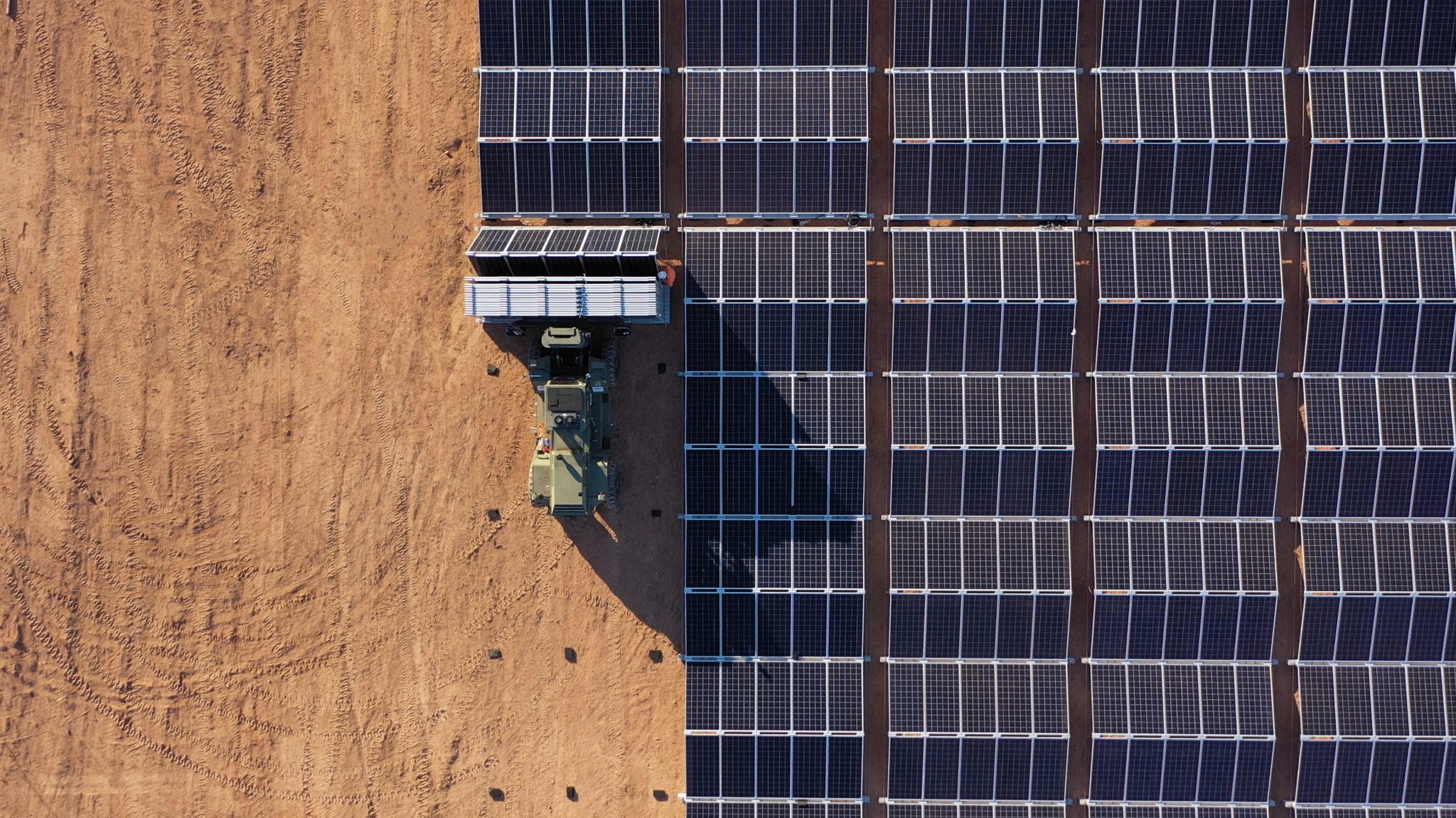 Aerial view of a solar farm under construction, showing a vehicle and a worker installing solar panels on a grid next to a dirt area.