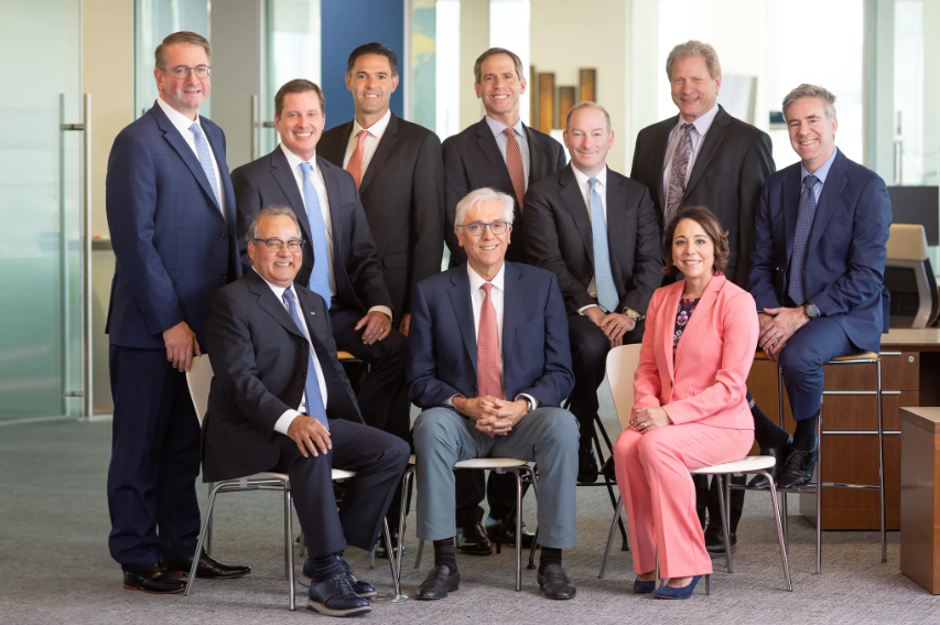 Group of nine men and one woman in business attire posing together in a modern office. Some are seated on chairs, while others stand behind them, all smiling at the camera. The office has large windows and contemporary furniture in the background.