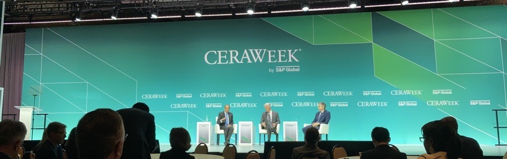 Three people are seated on a stage at CERAWeek by S&P Global. The stage backdrop is teal with geometric patterns and the CERAWeek logo. The audience is visible in the foreground.