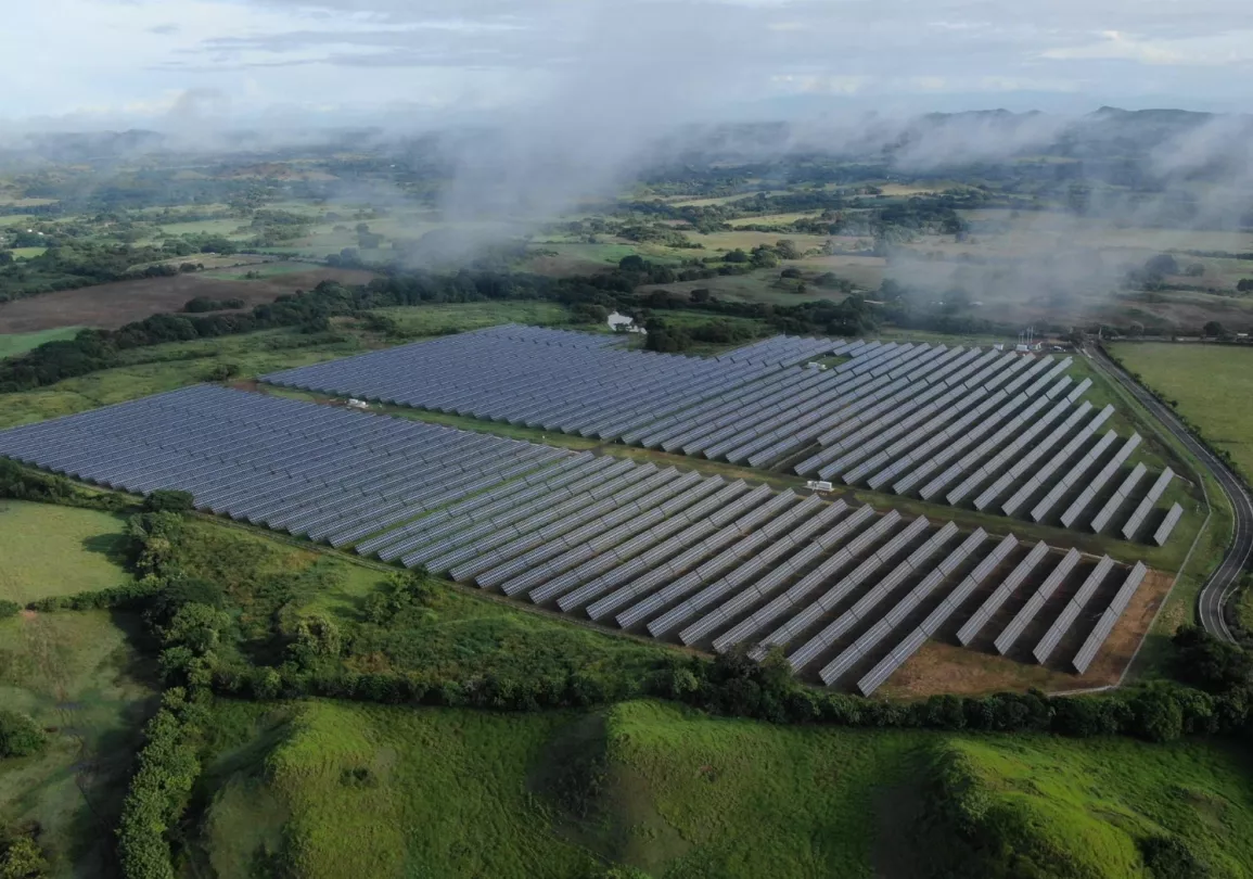 Aerial view of a large solar farm with rows of solar panels set in a green landscape. Misty hills and fields extend into the distance under a cloudy sky.