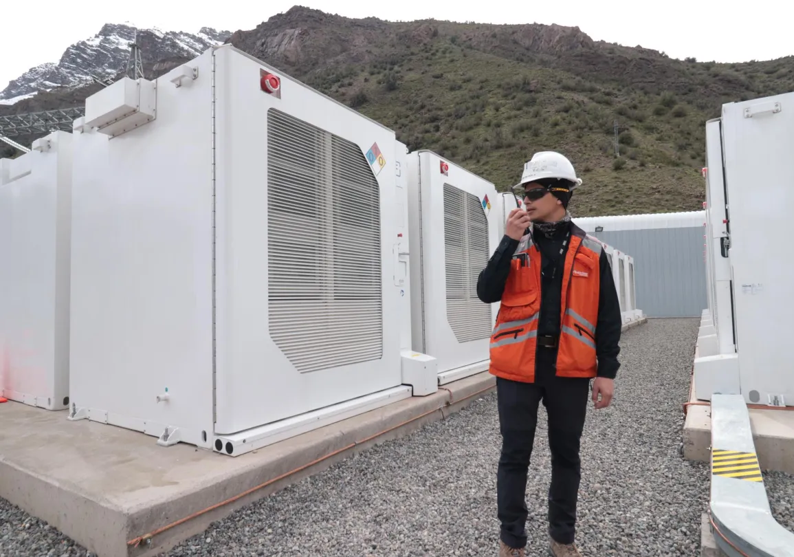 A worker in an orange safety vest and hard hat uses a walkie-talkie near large white industrial units. Snow-capped mountains are visible in the background.