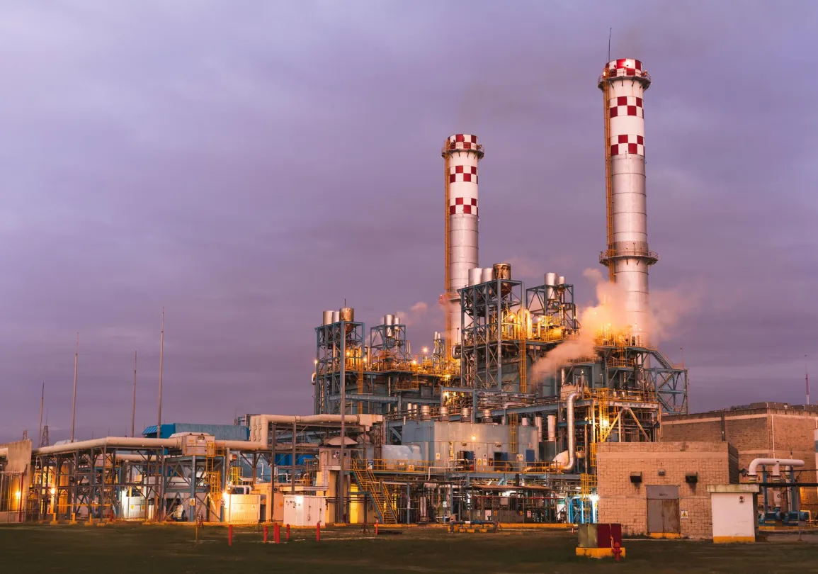 Industrial facility with two tall chimneys emitting steam, surrounded by metal structures and pipes, illuminated with lights against a cloudy sky at dusk.