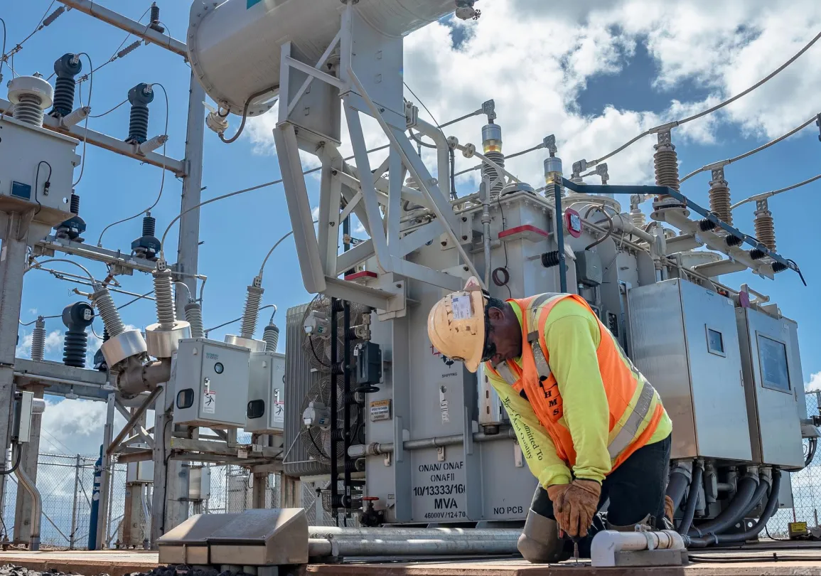 A worker in a hard hat and safety vest kneels at a power substation, surrounded by electrical equipment and transformers under a partly cloudy sky.