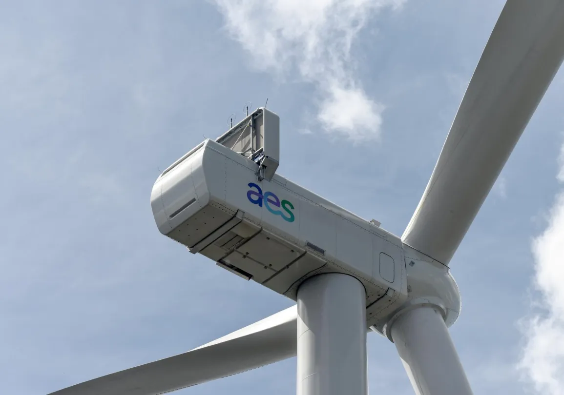 Close-up view of a wind turbine with a cloudy sky in the background. The turbine has the letters 'aes' on its nacelle.