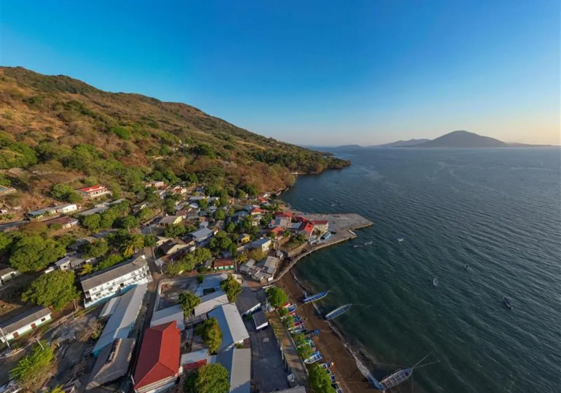 Aerial view of a coastal village with colorful houses nestled between a hillside and a body of water. Several small boats are docked along the shoreline.