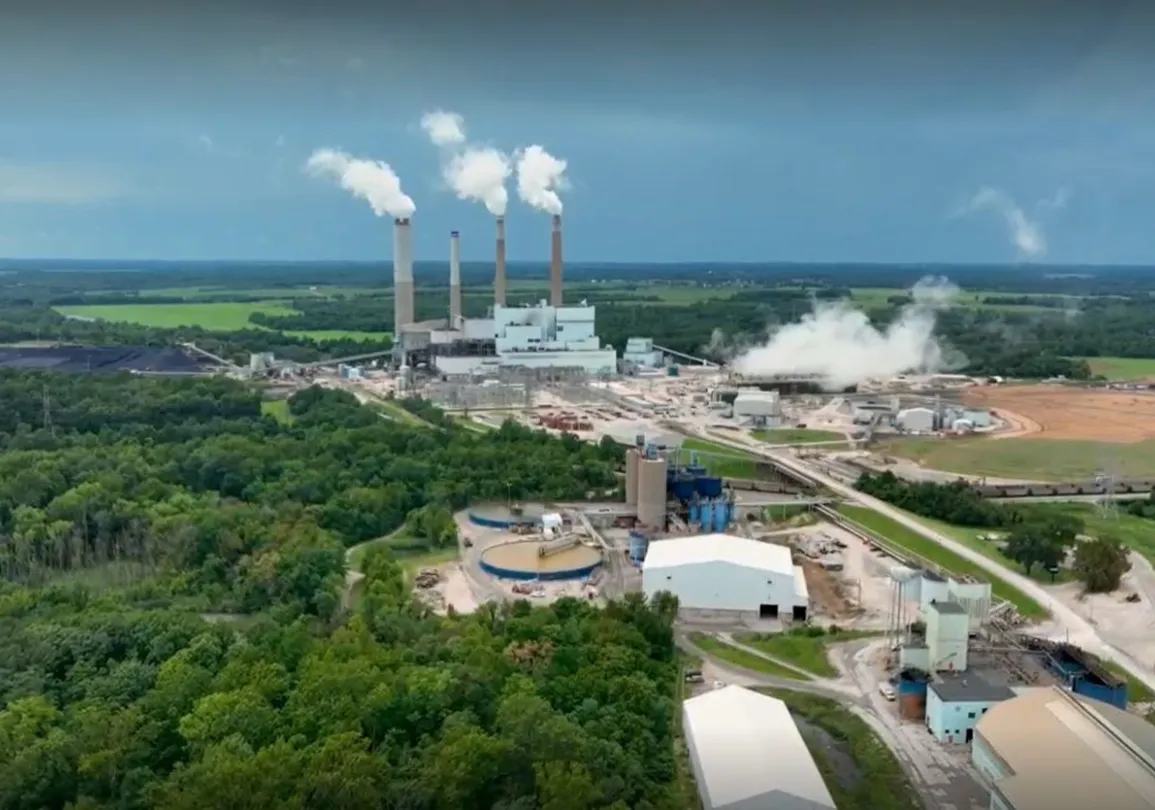 Aerial view of a large industrial plant with multiple chimneys emitting white smoke. The facility is surrounded by green fields and dense forested areas. Rail tracks and several buildings are visible in the foreground. The sky is overcast, suggesting a cloudy day.