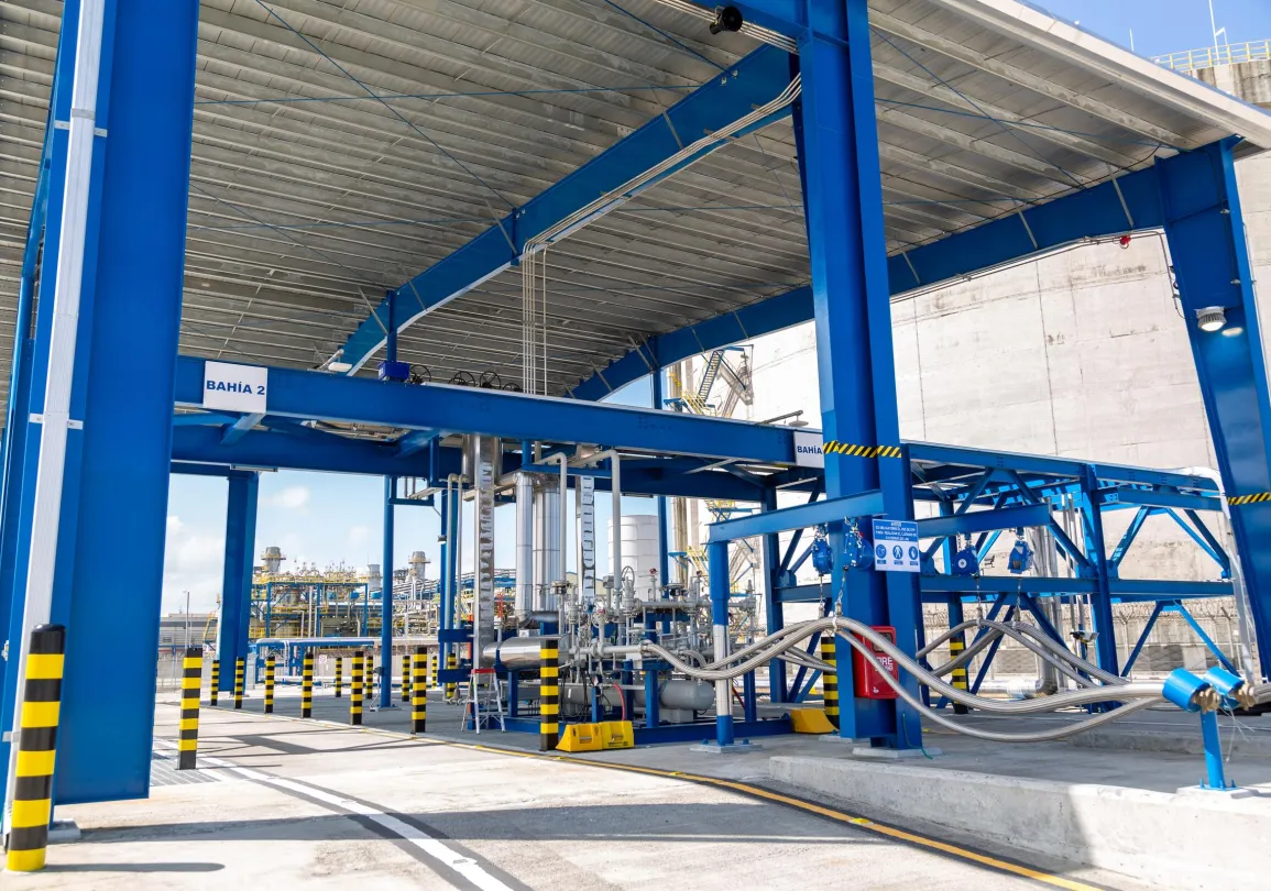 Industrial facility with blue metal framework and pipes, labeled 'BAHÍA 2'. The area has yellow and black striped barriers and safety signs. A large concrete wall is visible in the background.