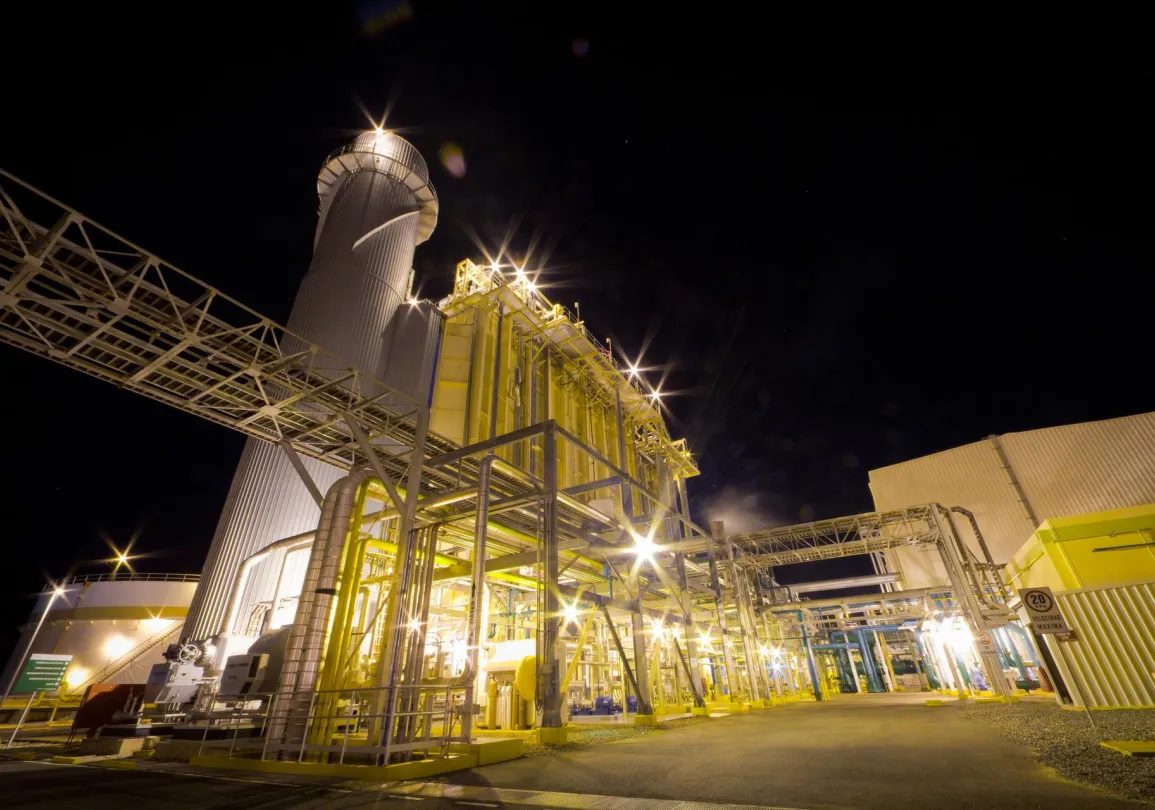 A brightly lit industrial facility at night, featuring a tall cylindrical tower and a network of metal structures and pipes. The lights create starburst effects against the dark sky, illuminating the yellow and metallic surfaces of the buildings and equipment.