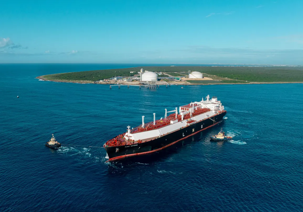Aerial view of a large LNG tanker being guided by two tugboats in a blue sea. The tanker is approaching an island with industrial facilities and storage tanks. The sky is clear and the water is calm.