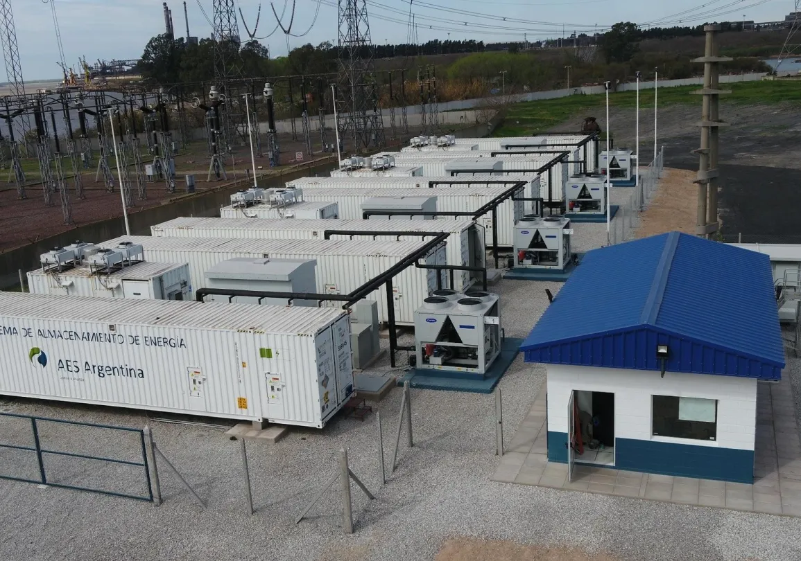 Aerial view of an energy storage facility with multiple white container units labeled 'AES Argentina' and a small building with a blue roof. Electrical infrastructure and power lines are visible in the background.