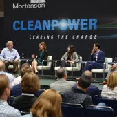 A panel of five people seated on a stage at a CleanPower event, with an audience in the foreground. The backdrop displays the words 'CleanPower Leading the Charge.'