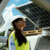 A person wearing a hard hat and safety vest inspects solar panels under a clear blue sky.