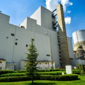 A large industrial building with a corrugated metal exterior, surrounded by green shrubs and trees. A tall smokestack and cooling tower are visible in the background. The sky is clear with a few clouds.
