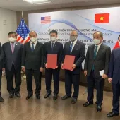 A group of seven men in suits and masks stand in a room with American and Vietnamese flags. Two men in the center hold red folders. A banner behind them reads 'Handover Ceremony of Commercial Agreements' in English and Vietnamese.
