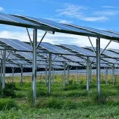 A field of solar panels mounted on metal frames, set against a blue sky with scattered clouds. The panels are arranged in rows over a grassy area.