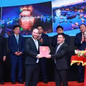 Two men in suits stand on a stage, holding a red document together. Behind them, several other people in suits are clapping. A woman in a red dress holds a floral arrangement on a tray. The background features images of colorful cityscapes and fireworks.