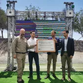 Four men stand on a green platform outdoors, holding a plaque that reads 'AES Chile destaca el compromiso de Sierra Gorda SCM'. Behind them is a large screen and metal framework. Trees and a clear blue sky are visible in the background.