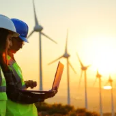 Two workers wearing hard hats and reflective vests stand in front of wind turbines at sunset. One holds a laptop, while the other looks on. The sky is a warm gradient of colors, and the wind turbines are silhouetted against the light.