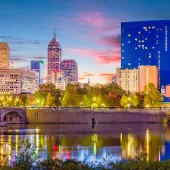 A city skyline at dusk with tall buildings, including a prominent blue glass structure, reflecting in a calm river. A bridge spans the river, and trees line the riverbank, illuminated by streetlights. The sky is a mix of blue and pink hues.