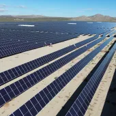 Aerial view of a large solar farm with rows of solar panels stretching across a desert landscape under a clear blue sky.