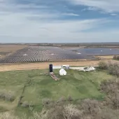 Aerial view of a large solar farm with rows of solar panels on a vast field. In the foreground, there is a small farm with a barn and silo surrounded by green fields and trees. The sky is partly cloudy.