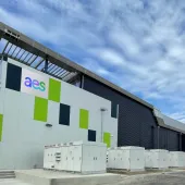 A modern industrial building with a sign displaying 'aes' on the facade. The building has a sleek design with green and dark panels. Several large electrical cabinets are positioned in front of it. The sky is partly cloudy.