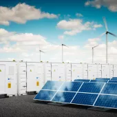 Image of an energy storage facility featuring rows of white containers labeled 'Energy Storage' with a blue lightning bolt symbol. In the foreground, there are several solar panels, and in the background, wind turbines are visible under a partly cloudy sky.