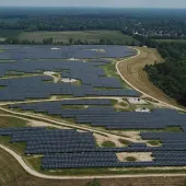Aerial view of a large solar farm with numerous solar panels arranged in rows across a vast green landscape. A small building and several vehicles are visible in the foreground. Trees and open fields surround the area, with a river running nearby.