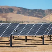 Rows of solar panels in a dry landscape with hills in the background under a clear blue sky.