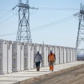 Two workers walk along a row of large white battery storage units with power lines and towers in the background under a clear blue sky.
