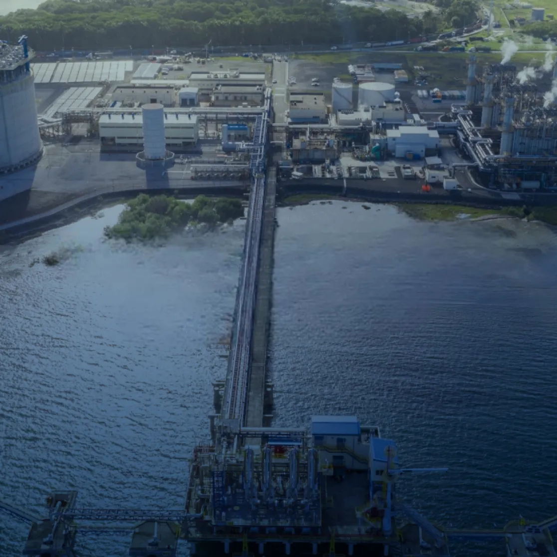 Aerial view of an industrial facility by the water, featuring large storage tanks, pipelines, and buildings. A long pier extends into the water, connecting the facility to the shoreline.