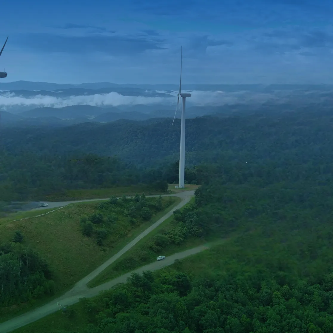 Aerial view of a wind farm with multiple wind turbines on a lush green hilltop, surrounded by dense forests and distant mountain ranges under a clear blue sky.