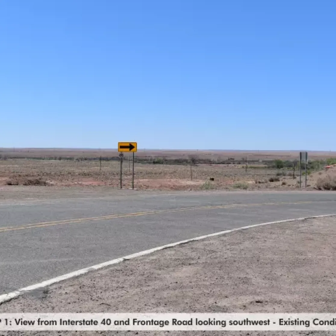 A view from Interstate 40 and Frontage Road looking southwest. The image shows a wide expanse of flat, arid land under a clear blue sky. A road curves in the foreground with a yellow arrow sign indicating a left turn. Sparse vegetation is visible in the distance.