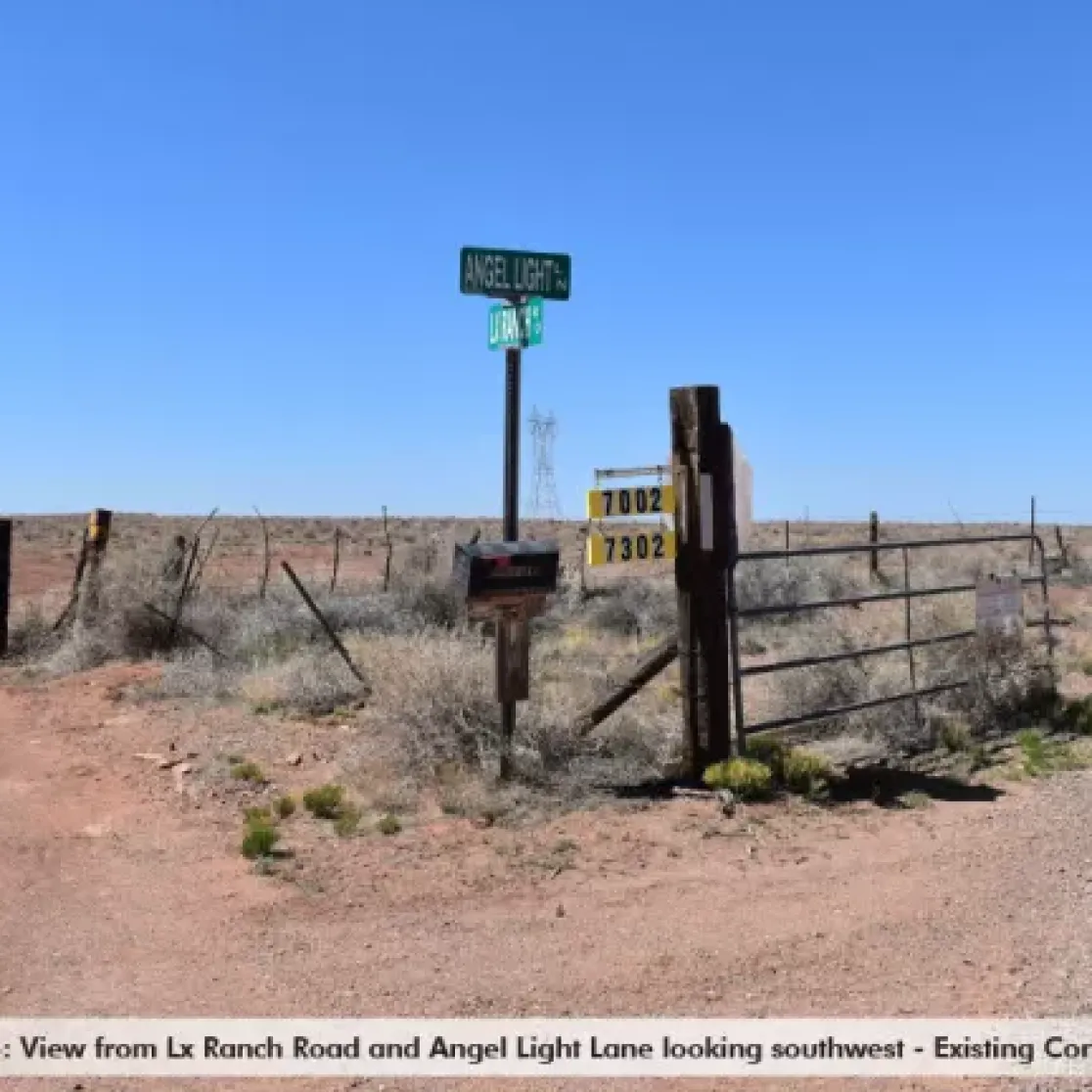 A rural intersection with a dirt road and a metal gate. Signs indicate 'Angel Light Ln' and 'Lx Ranch Rd'. The area is flat with sparse vegetation and a clear blue sky. A small post displays numbers 7002 and 7302.