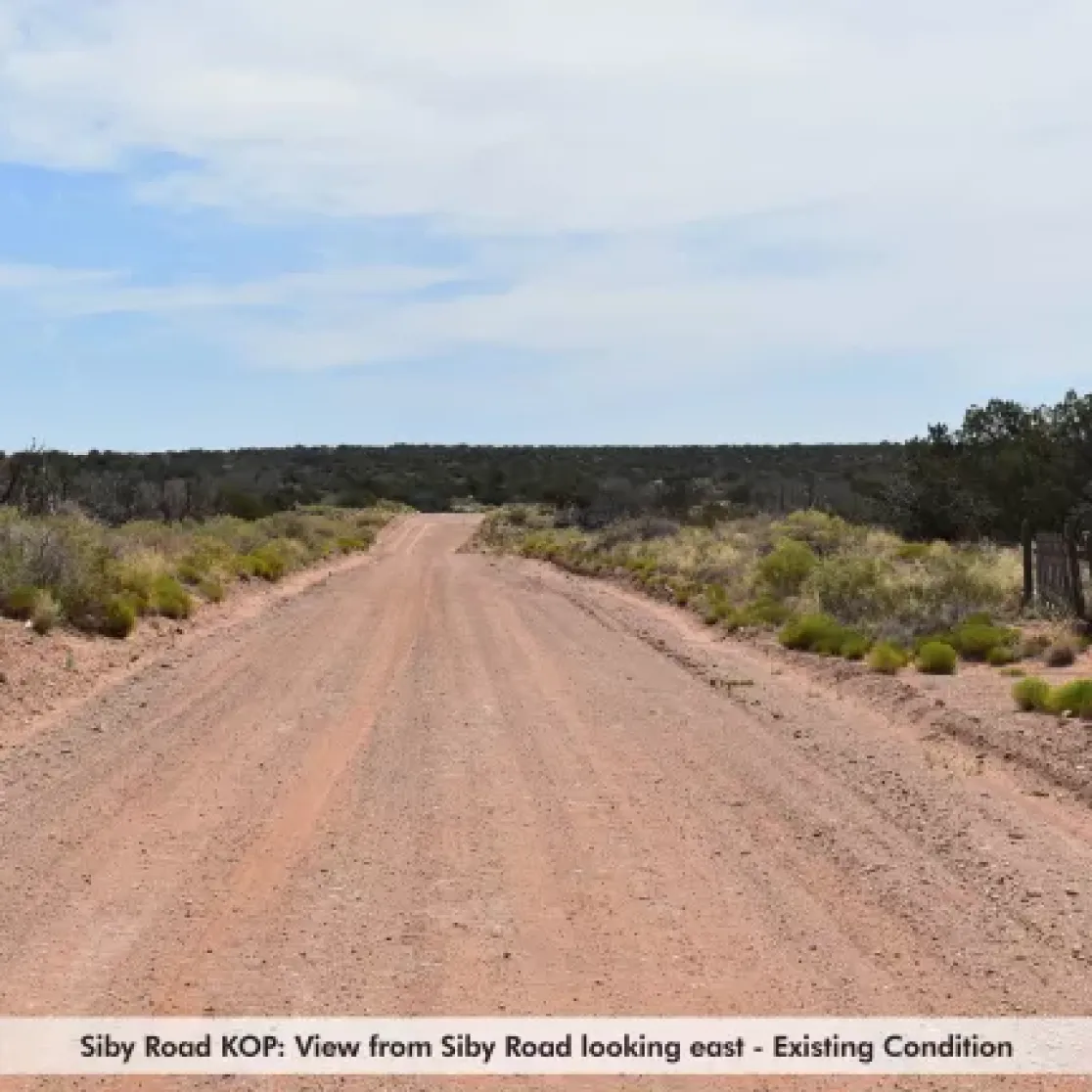 A dirt road stretches into the distance under a blue sky with scattered clouds. The road is flanked by sparse vegetation and low shrubs on both sides. The landscape is flat with a line of trees in the background. A caption at the bottom reads, 'Siby Road KOP: View from Siby Road looking east - Existing Condition.'