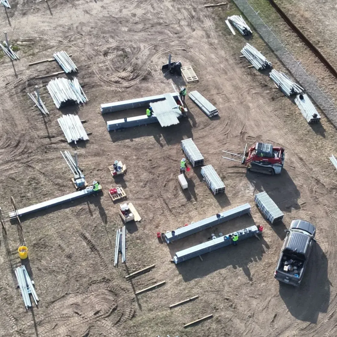 Aerial view of a construction site with scattered metal beams and materials on a dirt field. Workers and machinery are visible, including a red loader and a truck. A small white building with a curved roof is on the right, surrounded by grass.