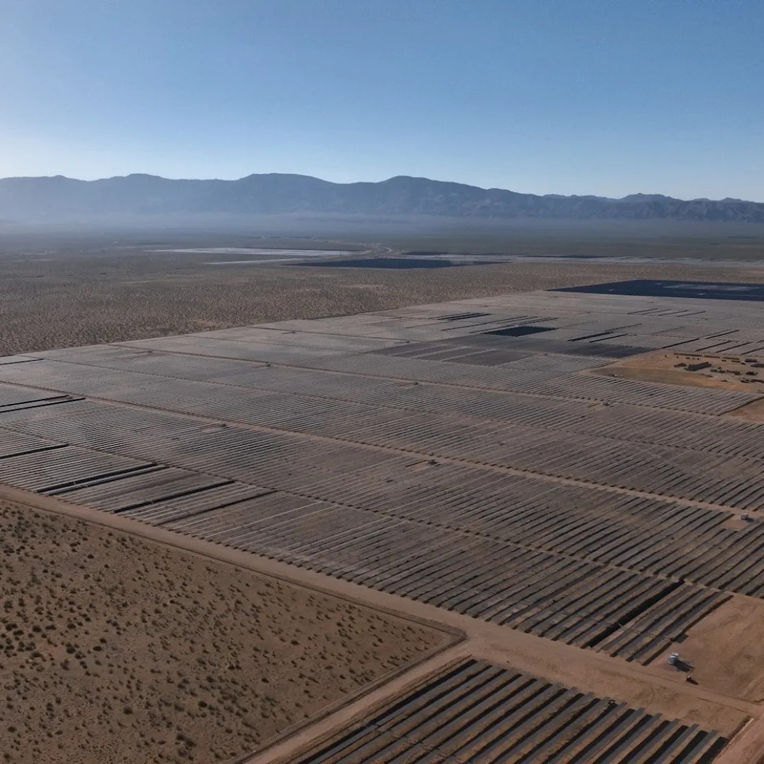 Aerial view of a large solar farm in a desert landscape. Rows of solar panels stretch across the sandy terrain. Mountains are visible in the background under a clear blue sky.