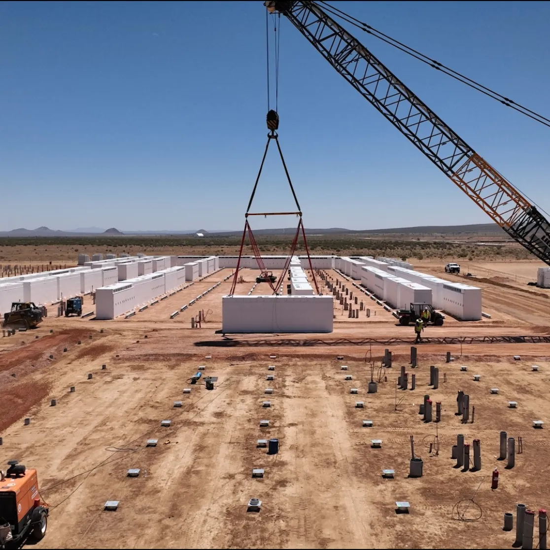 A construction site in a desert landscape with a large crane lifting a white rectangular structure. Rows of similar structures are aligned on the ground. Heavy machinery and workers are visible, with mountains in the distant background under a clear blue sky.