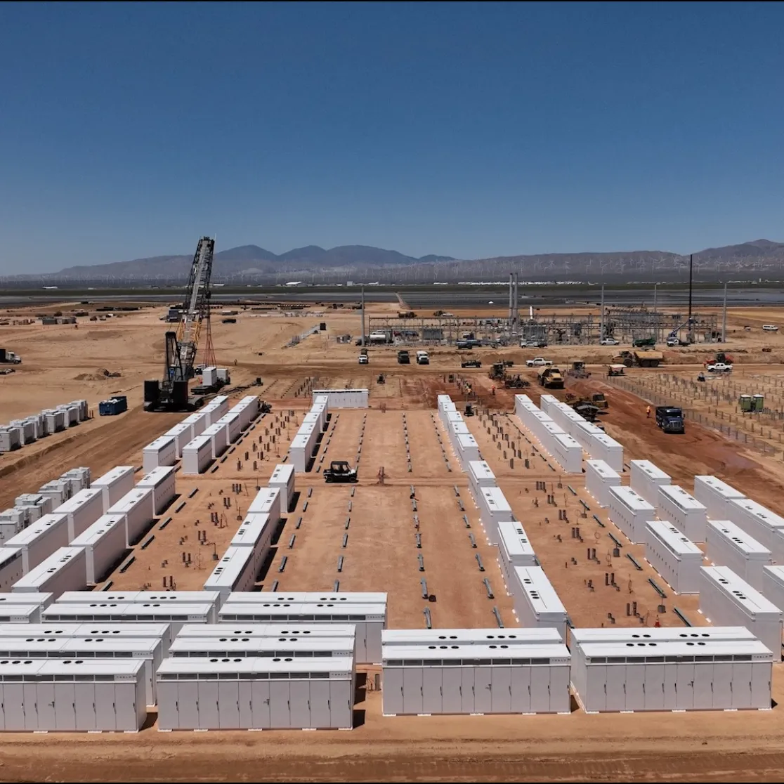 Aerial view of a construction site in a desert landscape with rows of white rectangular units, possibly battery storage, arranged in a grid pattern. A crane is positioned centrally, with various construction vehicles and equipment scattered around. Mountains are visible in the distant background under a clear blue sky.