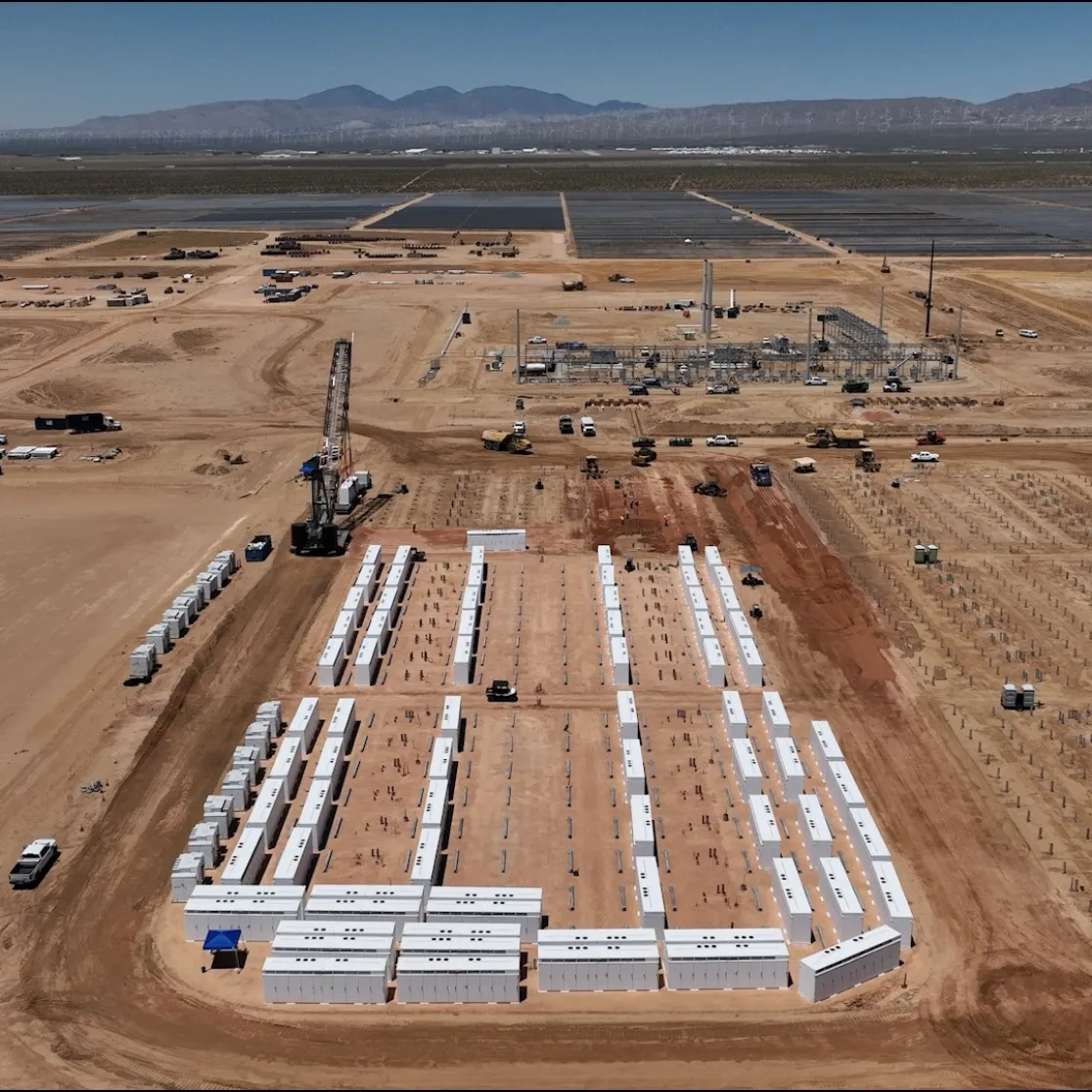 Aerial view of a large construction site in a desert landscape. Rows of white rectangular structures are neatly arranged in the foreground. Construction vehicles and equipment are scattered throughout the site. In the background, there are solar panels and mountains under a clear blue sky.